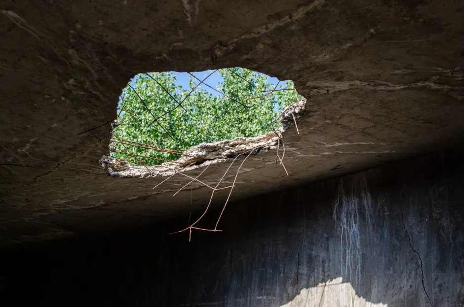 A large hole in a concrete structure provides a view of greenery and blue sky.